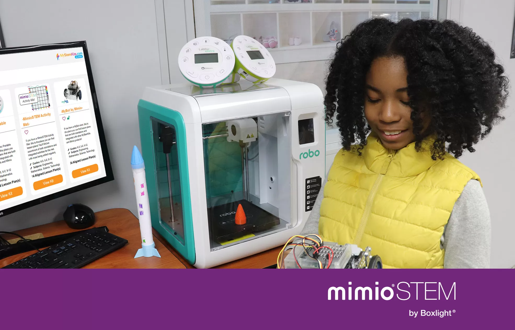 student holding an Educational Robotics System, standing infront of a 3D Printer and a screen displaying MyStemKits ​​​​​​STEM Curriculum