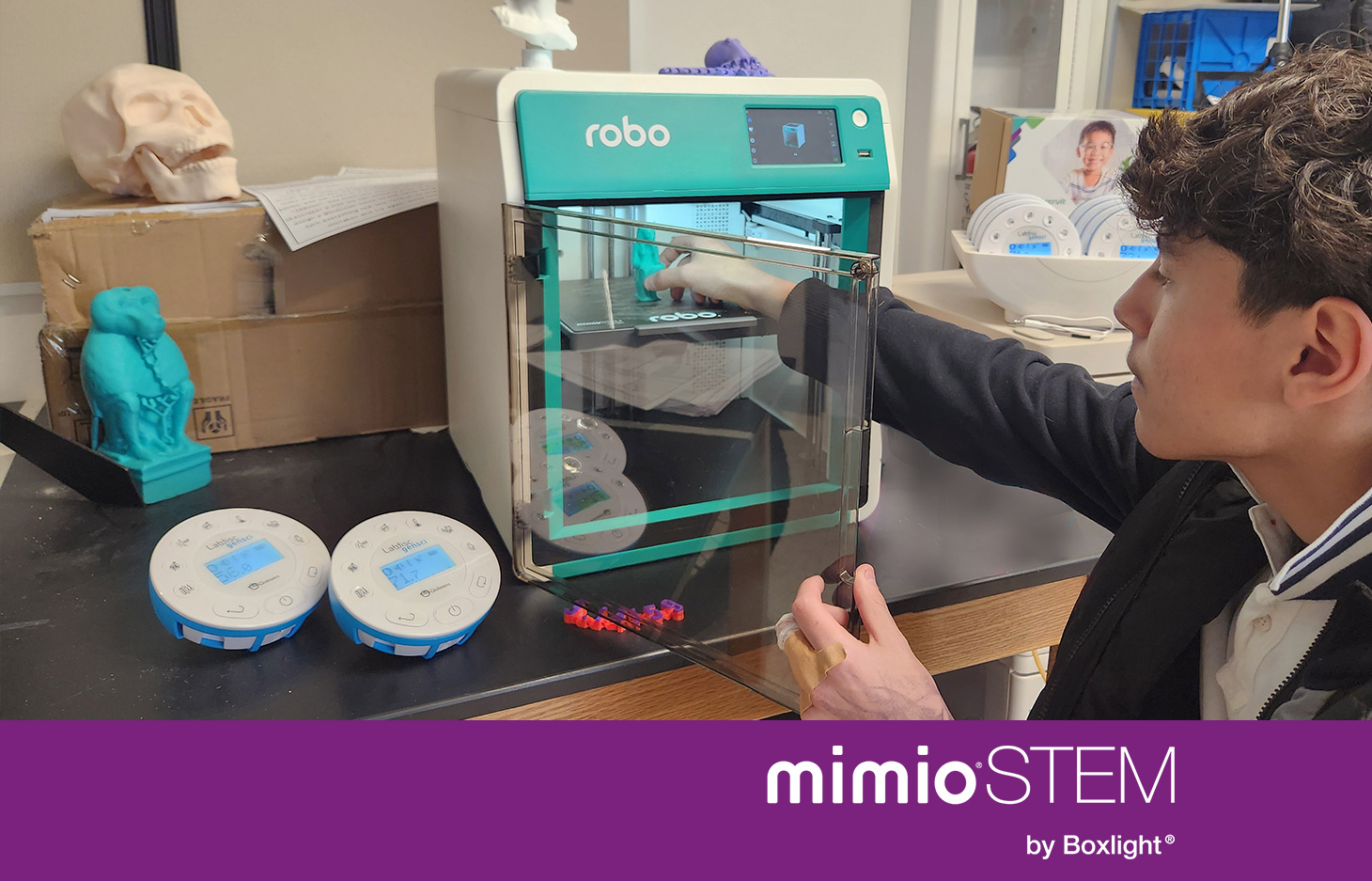 student holding an Educational Robotics System, standing infront of a 3D Printer and a screen displaying MyStemKits STEM Curriculum